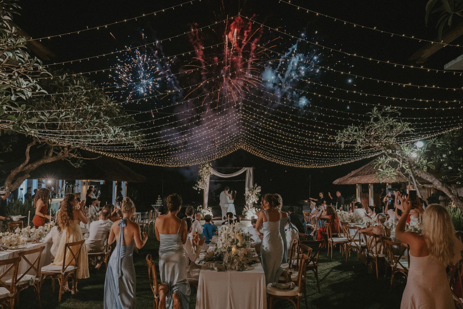 String lights illuminate an outdoor evening wedding reception with guests mingling under a starlit canopy.
