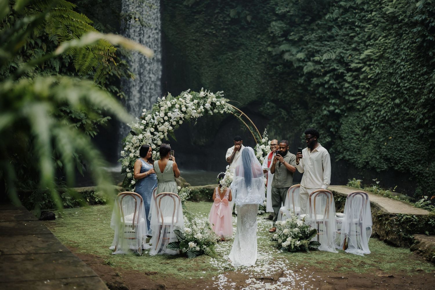 Outdoor wedding ceremony with floral arch and waterfall backdrop in a lush green garden setting.