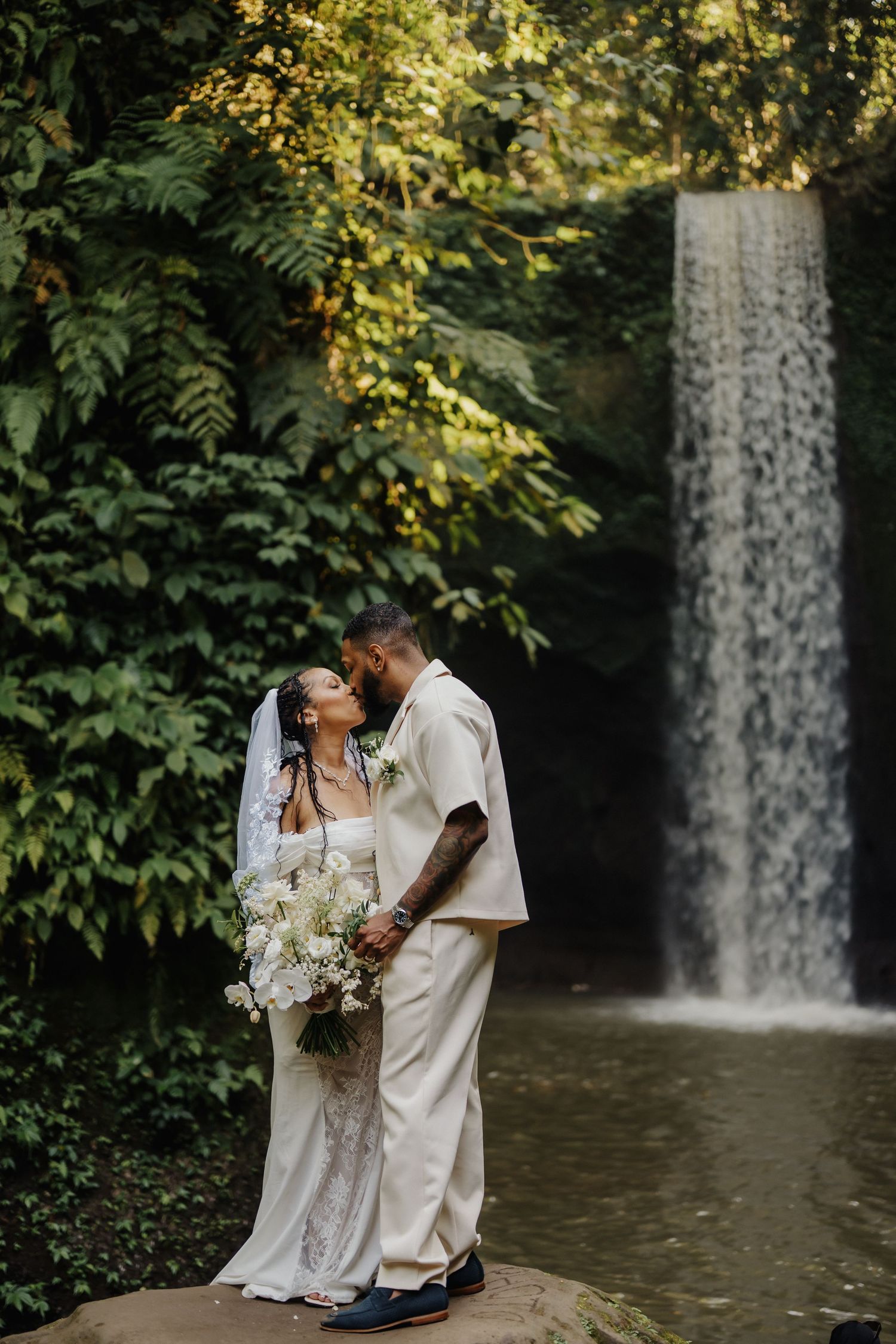 Bride and groom share a kiss in front of a lush waterfall, bride holding white bouquet in elegant lace gown.