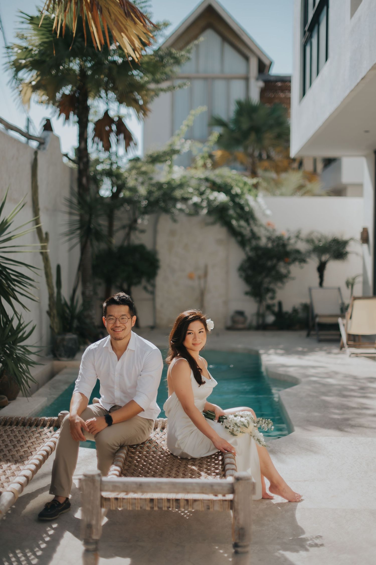 Couple sitting together on a wicker daybed by a pool in a tropical villa courtyard, man in white shirt, woman in white dress.