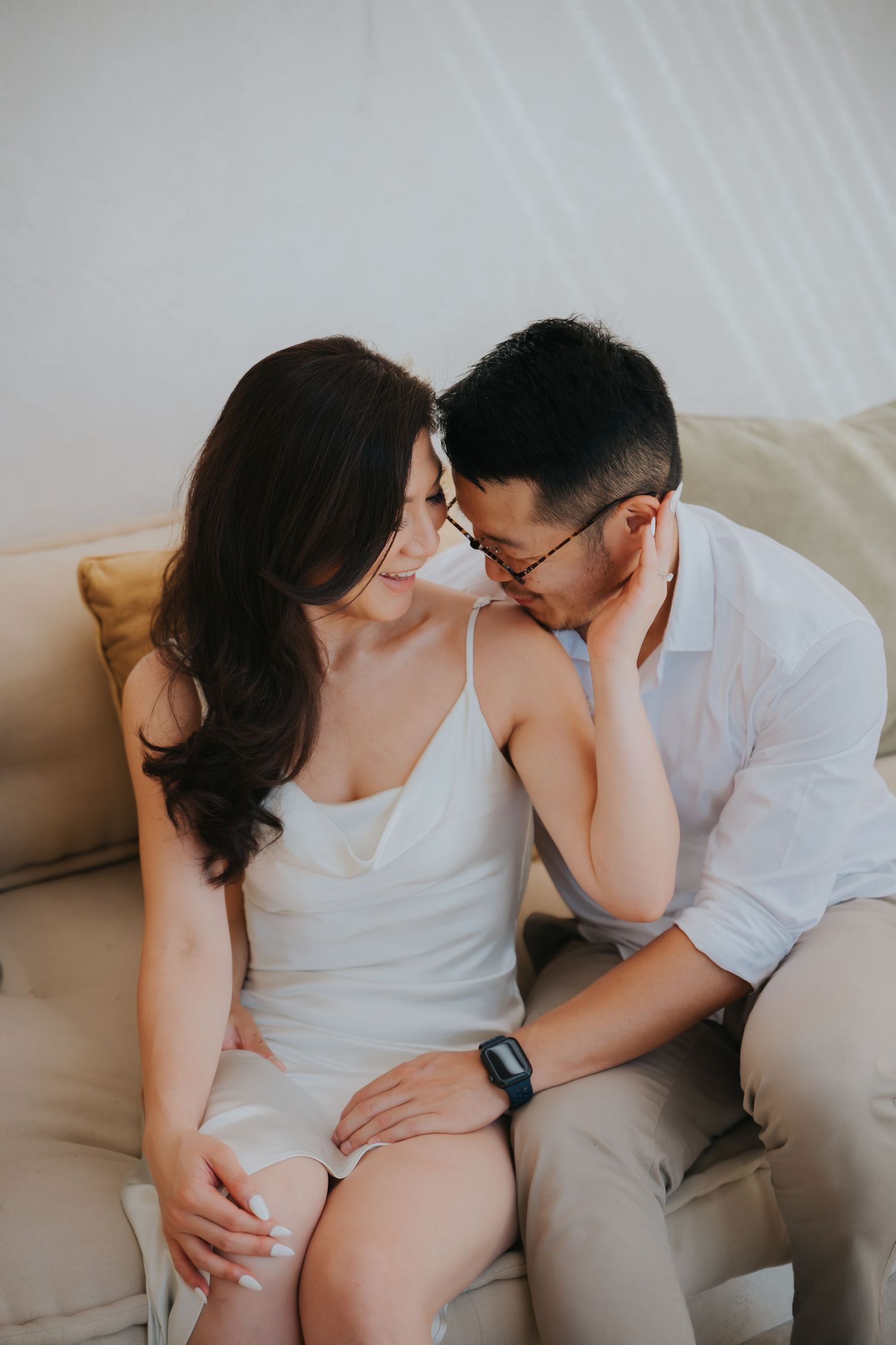 Romantic couple sharing an intimate moment on a couch, woman in white dress and man in white shirt with glasses.
