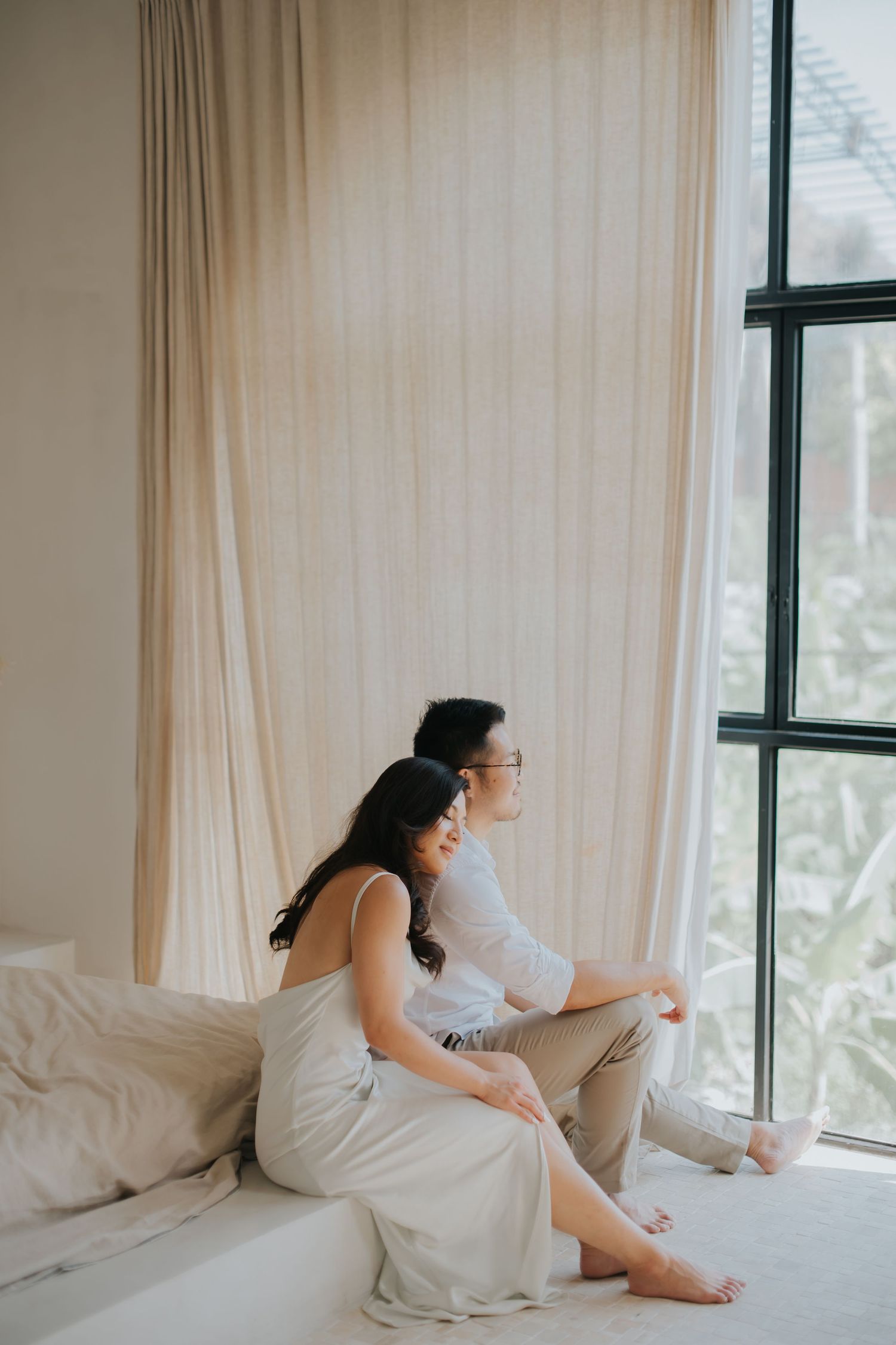 A couple sits together on a white couch by a large window, gazing outside in a bright, minimalist room.