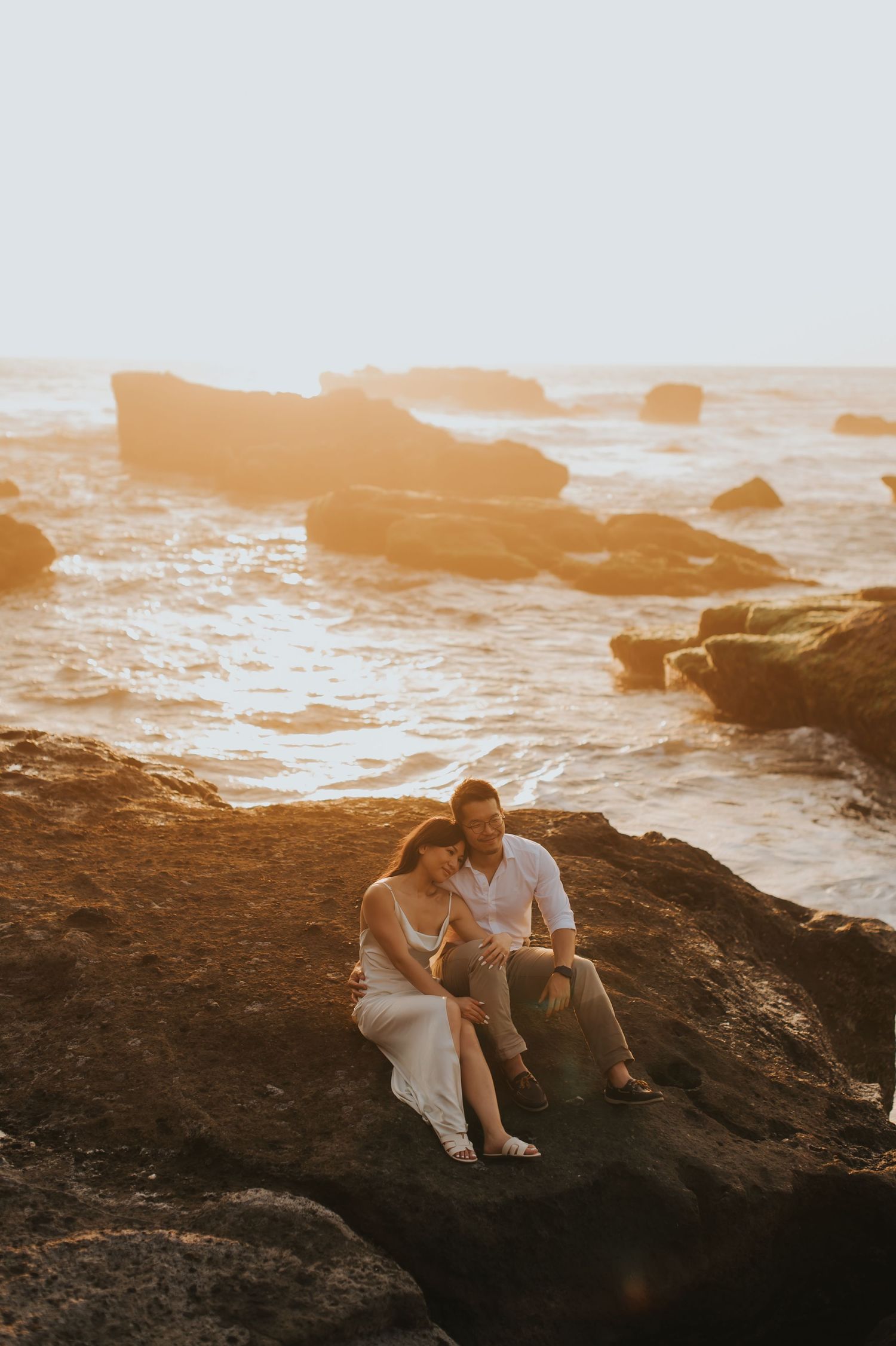 Couple sitting on rocky coastal cliffs during golden sunset, waves crashing on rocks behind them.