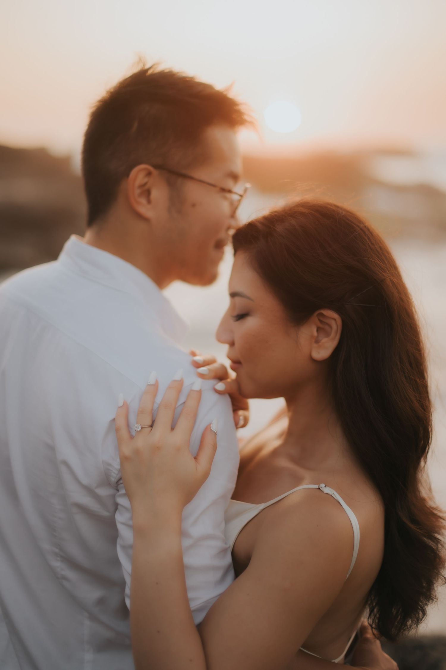 Couple sharing a romantic moment at golden hour sunset, woman wearing engagement ring, tender embrace outdoors.