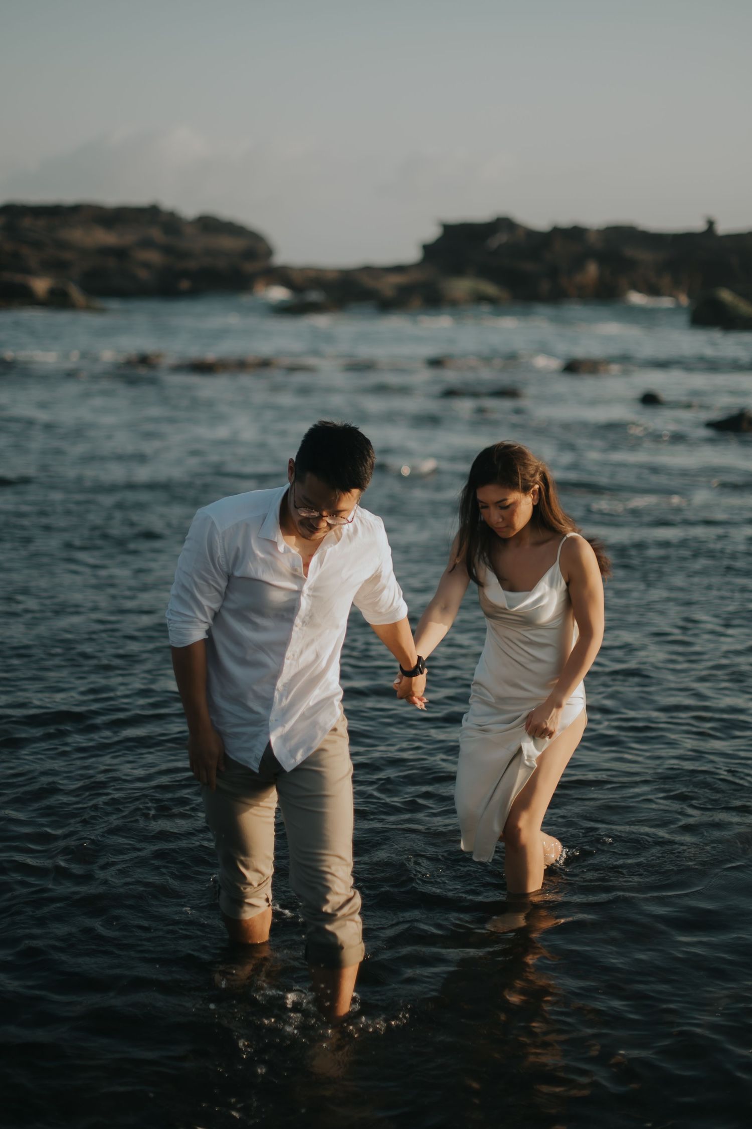 Couple holding hands wading through ocean water at sunset, dressed in white outfits on a rocky beach.