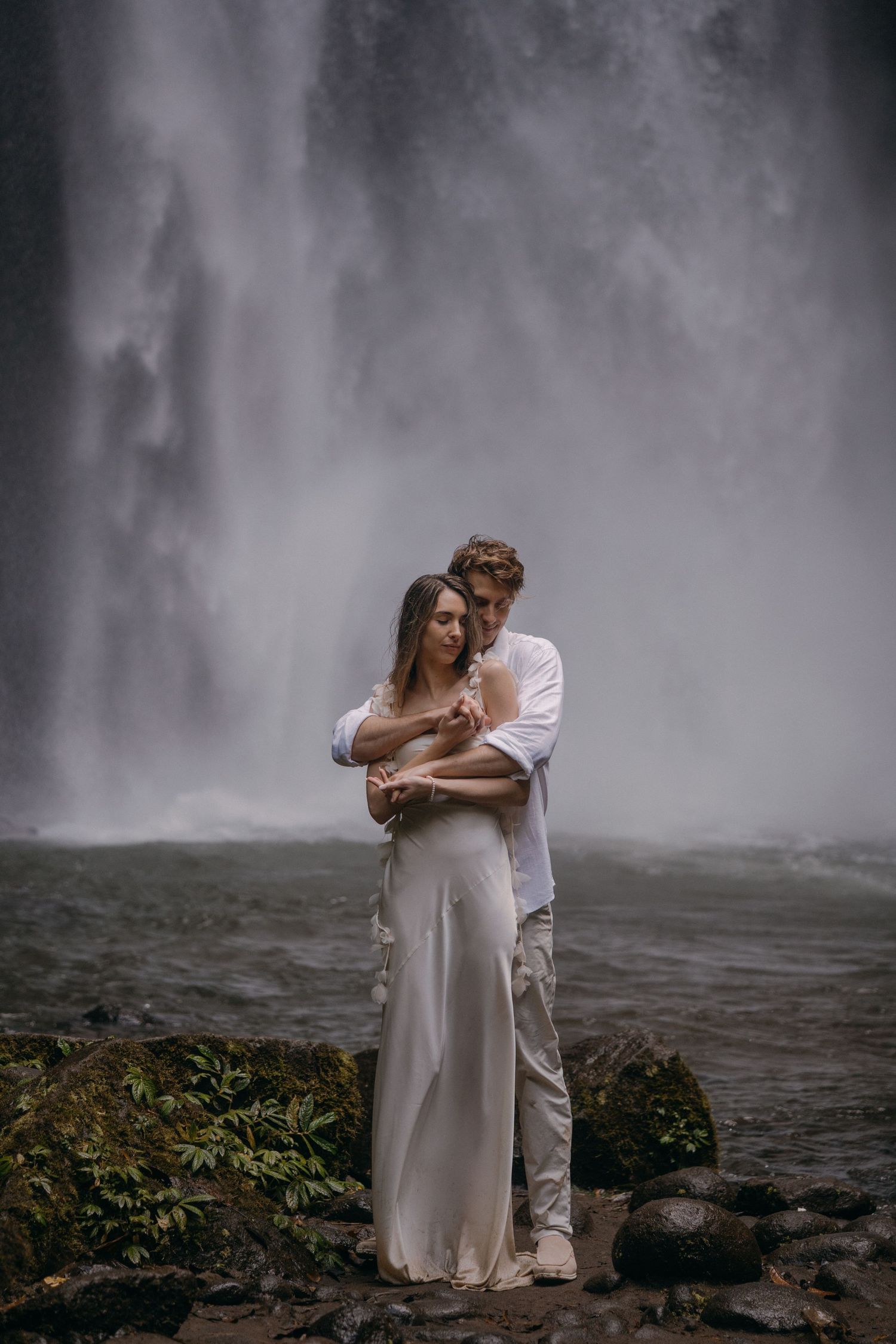 A romantic couple embraces in front of a misty waterfall, with the bride wearing a flowing white dress.