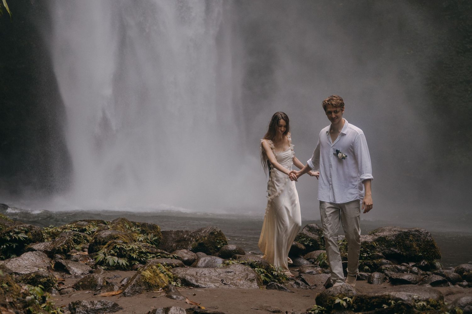A couple stands together on rocky terrain near a misty waterfall in an atmospheric outdoor setting.