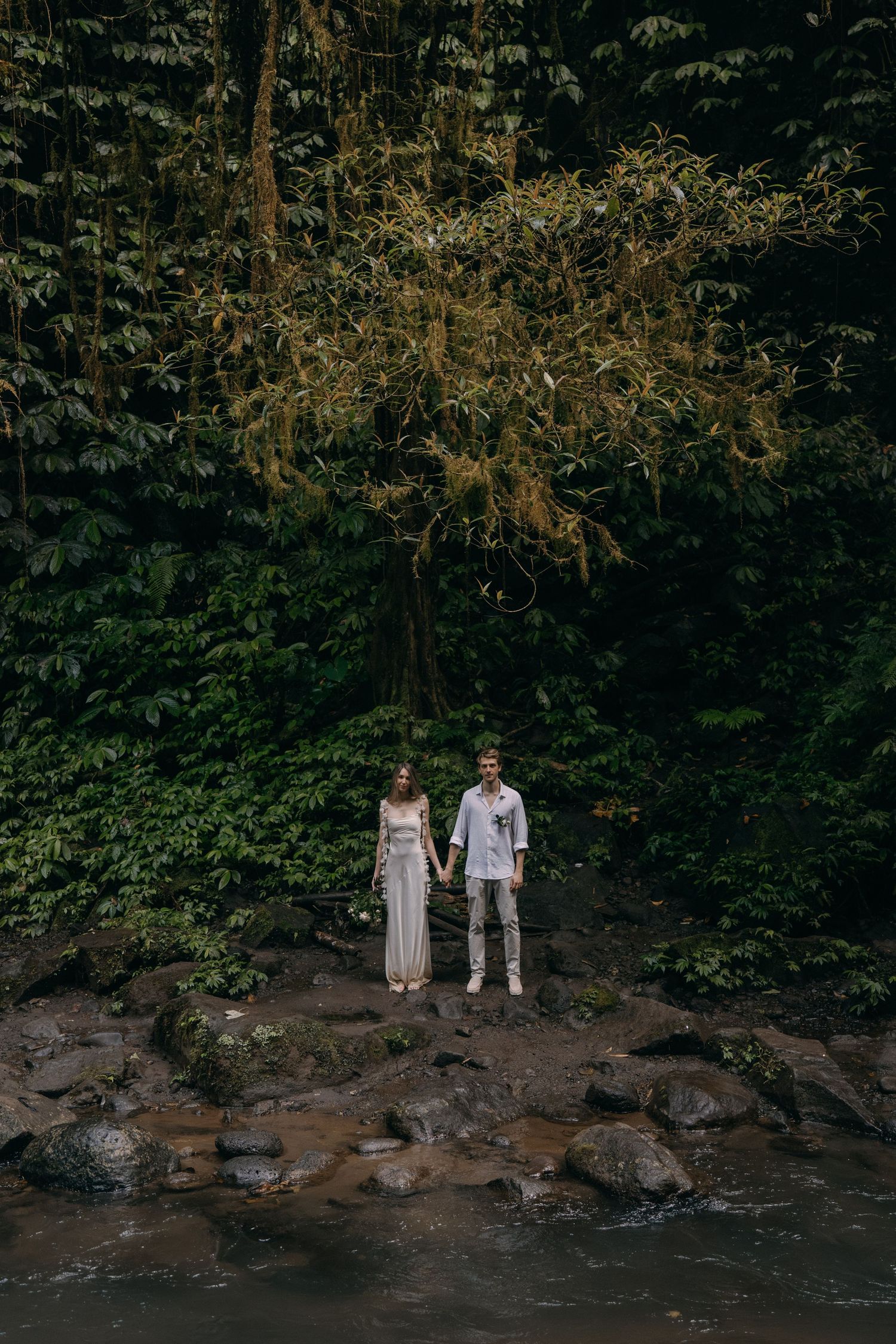 A couple in white attire stands beside a rocky stream under a glowing chandelier-like light fixture in a lush forest setting.