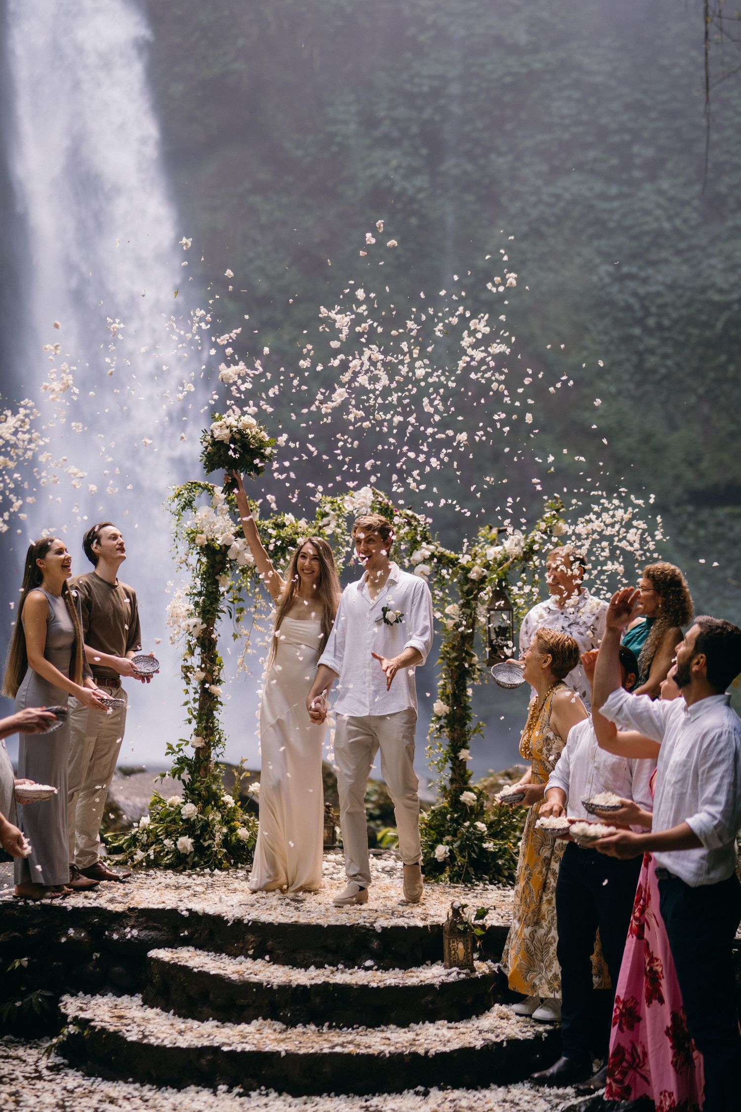 A wedding ceremony takes place on flower-adorned steps in front of a majestic waterfall as guests celebrate with confetti.