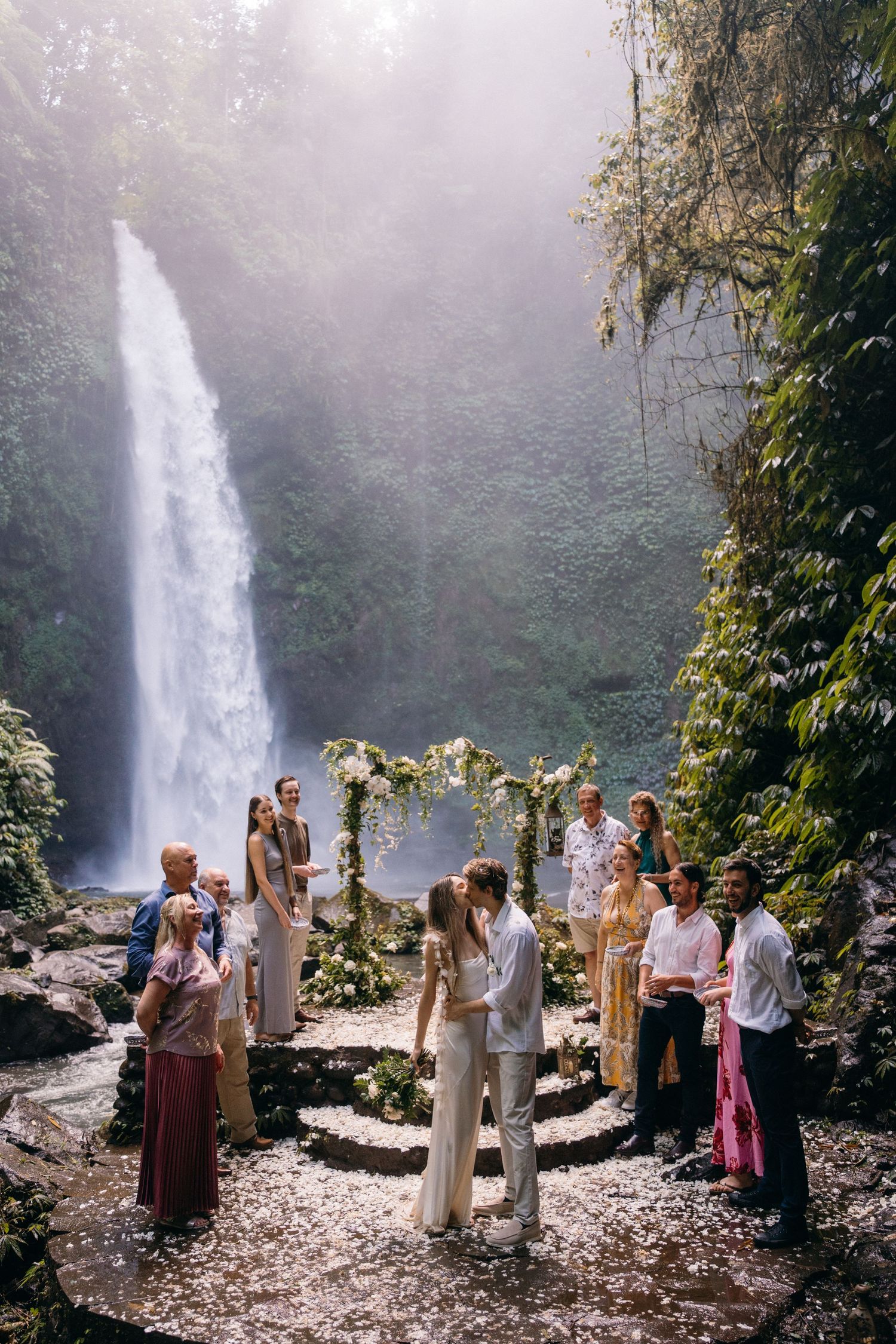 A wedding ceremony takes place in front of a majestic waterfall in a misty forest setting with floral arch and gathered guests.