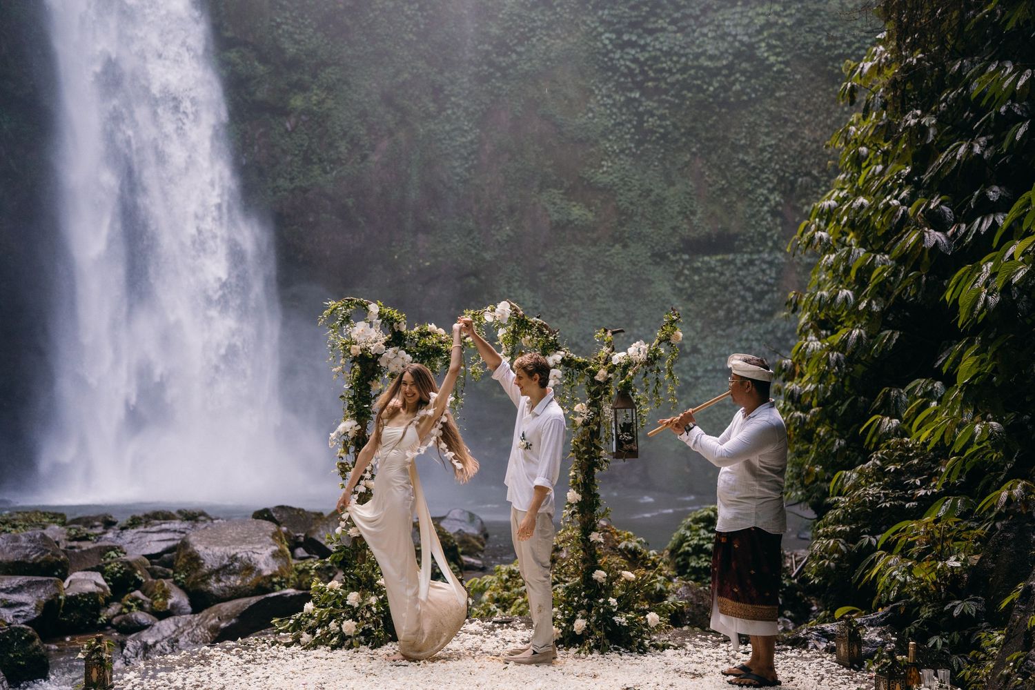 A wedding ceremony takes place under a floral arch against the backdrop of a majestic waterfall in nature.