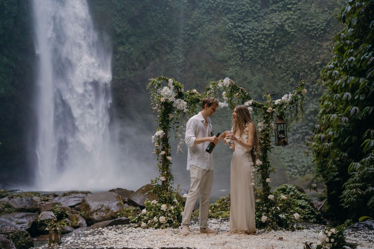 A romantic ceremony takes place in front of a majestic waterfall with a floral arch adorned with white blooms and greenery.