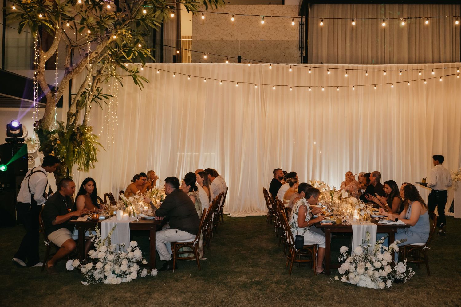 Wedding guests enjoy candlelit reception dinner at long tables beneath string lights and white draped backdrop at night.
