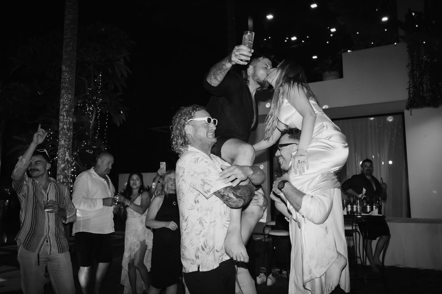 Bride is lifted by wedding guest as groom celebrates with raised bottle among dancing guests at night reception.