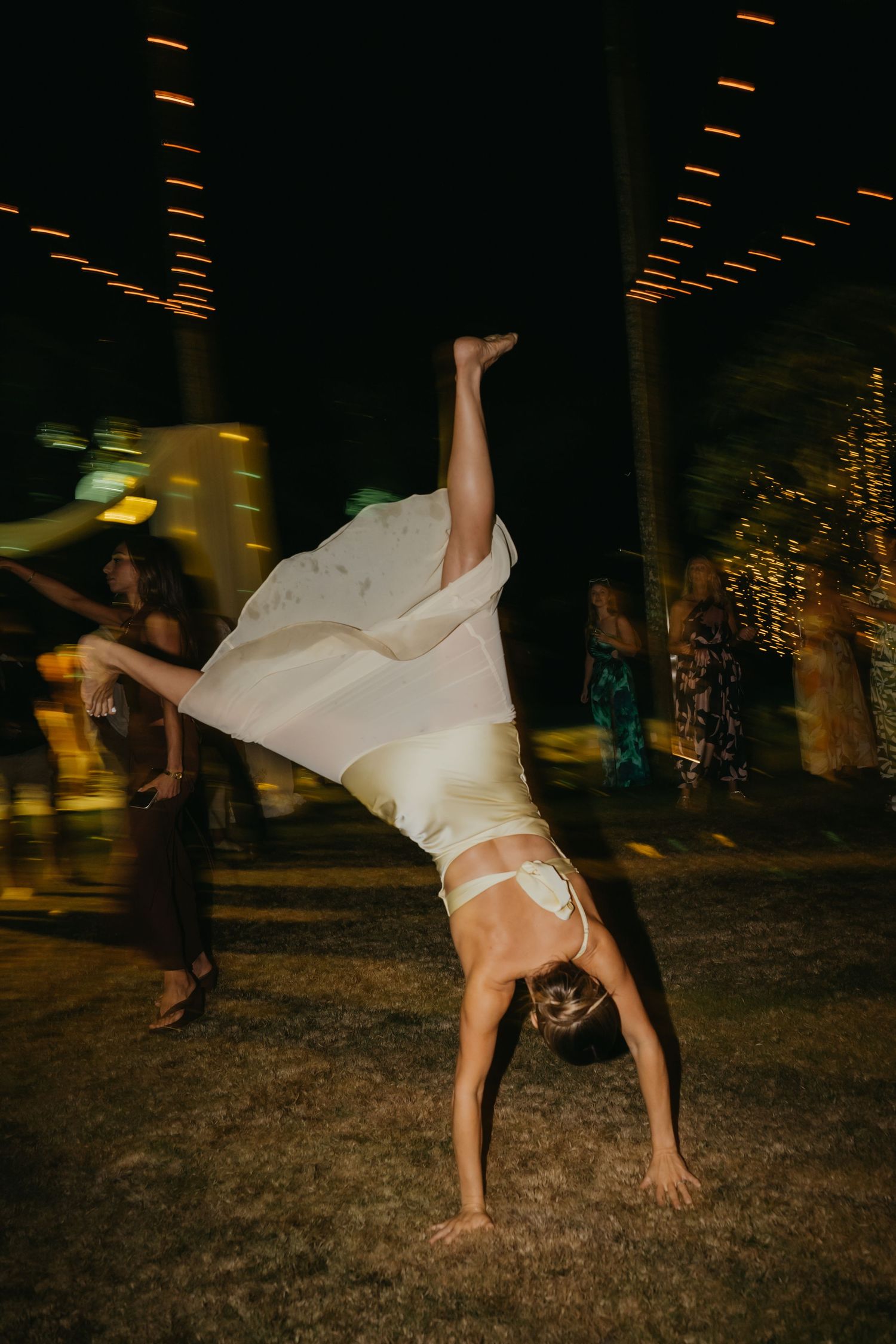 Bride performs a handstand in white satin gown during lively outdoor evening wedding reception.