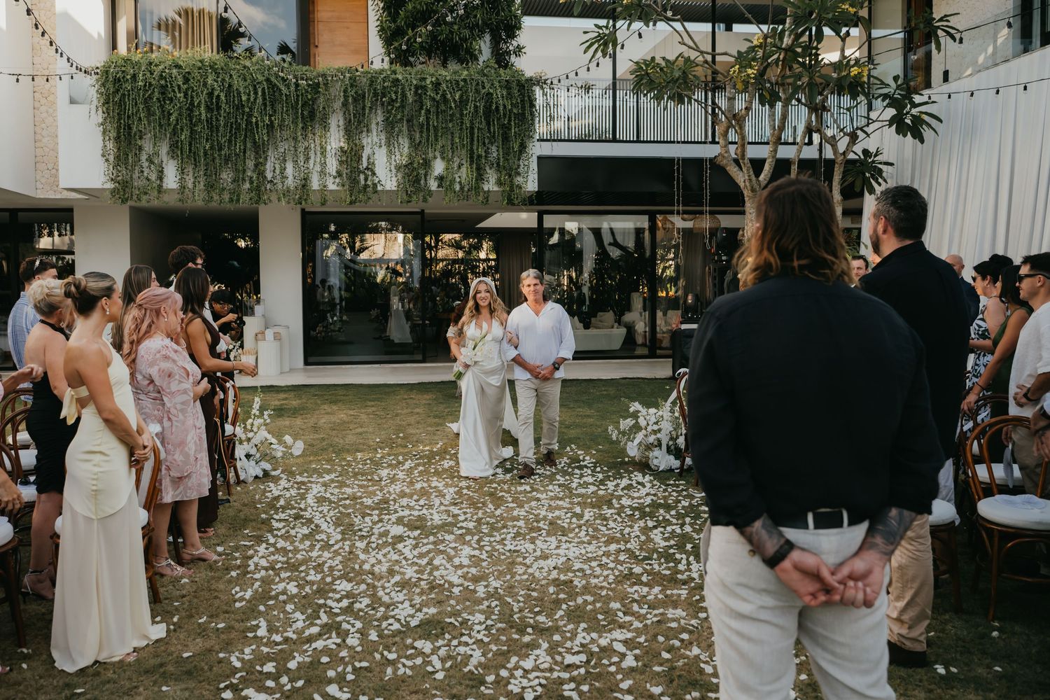 Couple stands at altar during outdoor wedding ceremony with guests seated along petal-covered aisle.