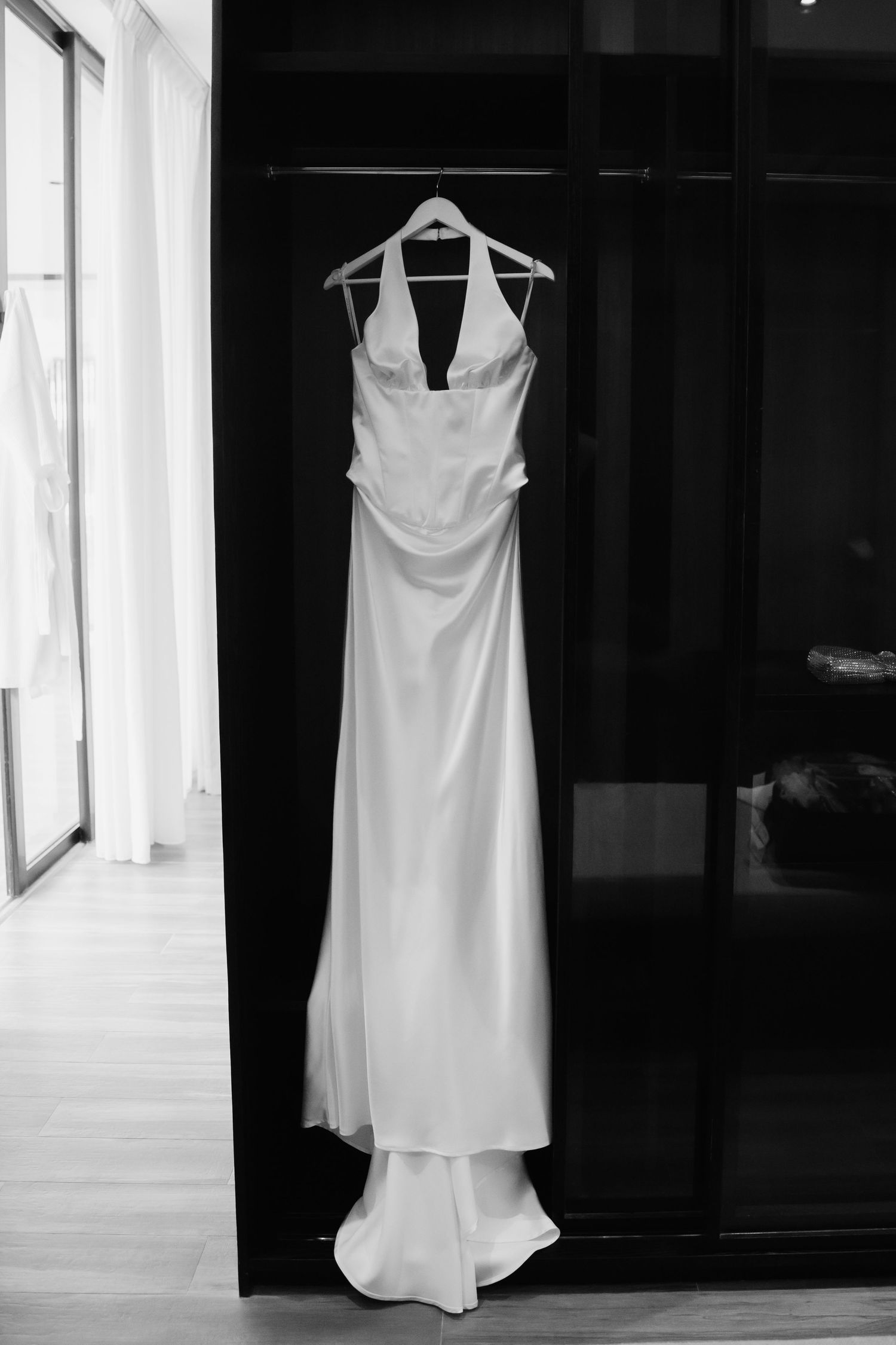 Sleek white satin halter wedding gown hanging on a wooden hanger against a dark wardrobe in black and white.