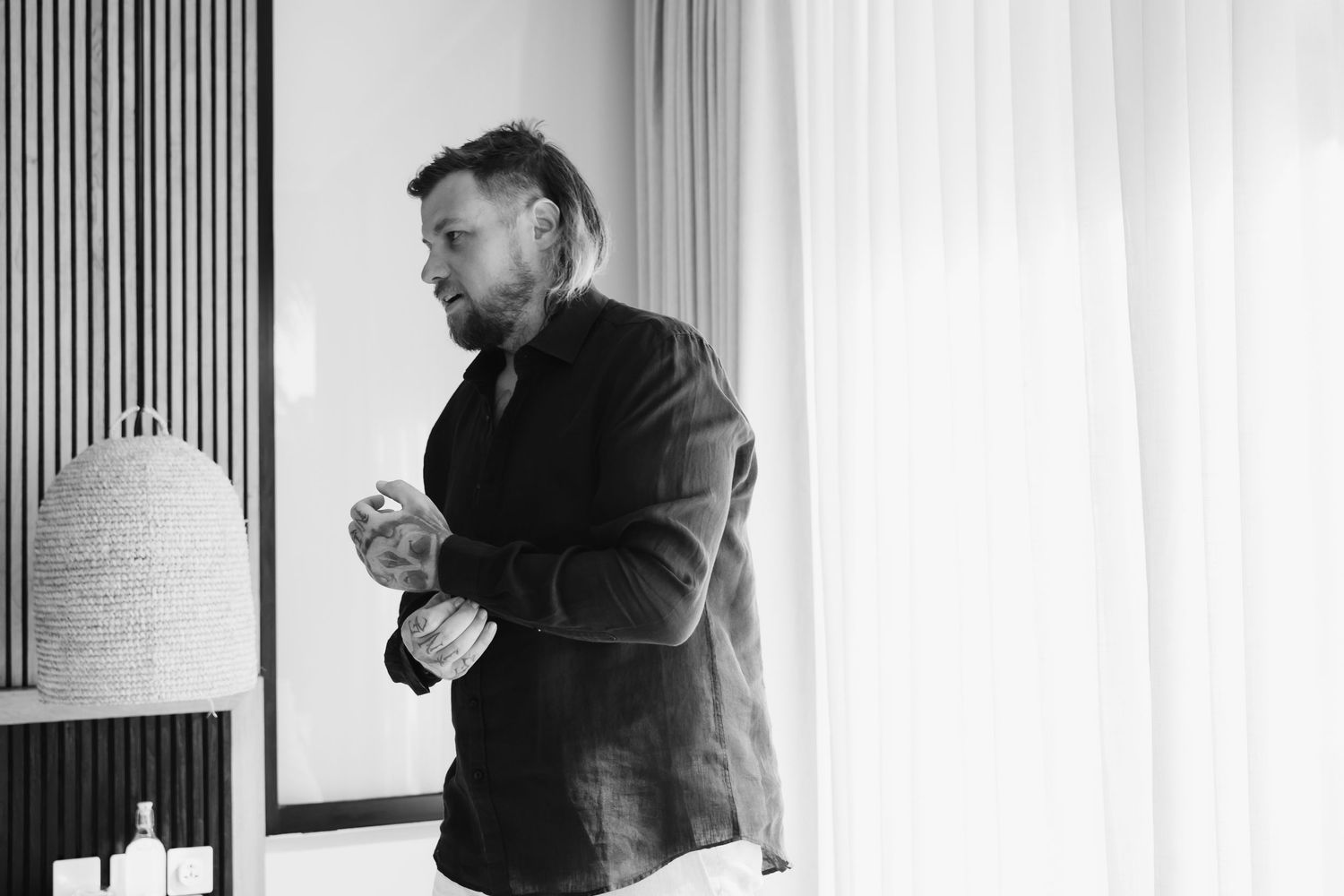 Groom in dark blazer adjusting cufflinks in bright room during wedding preparations.