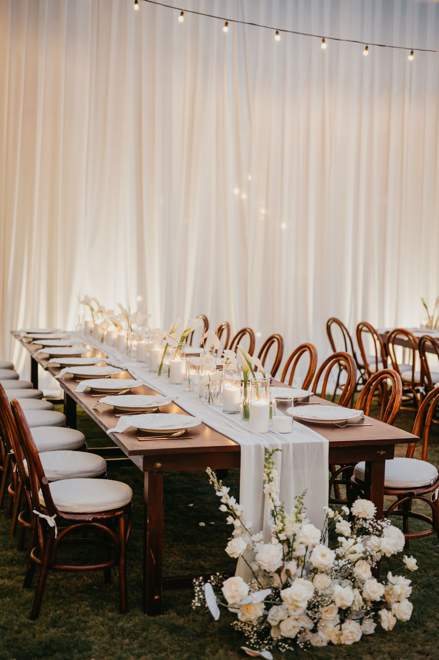 Elegant wedding reception with long wooden tables, white floral centerpieces, and string lights.