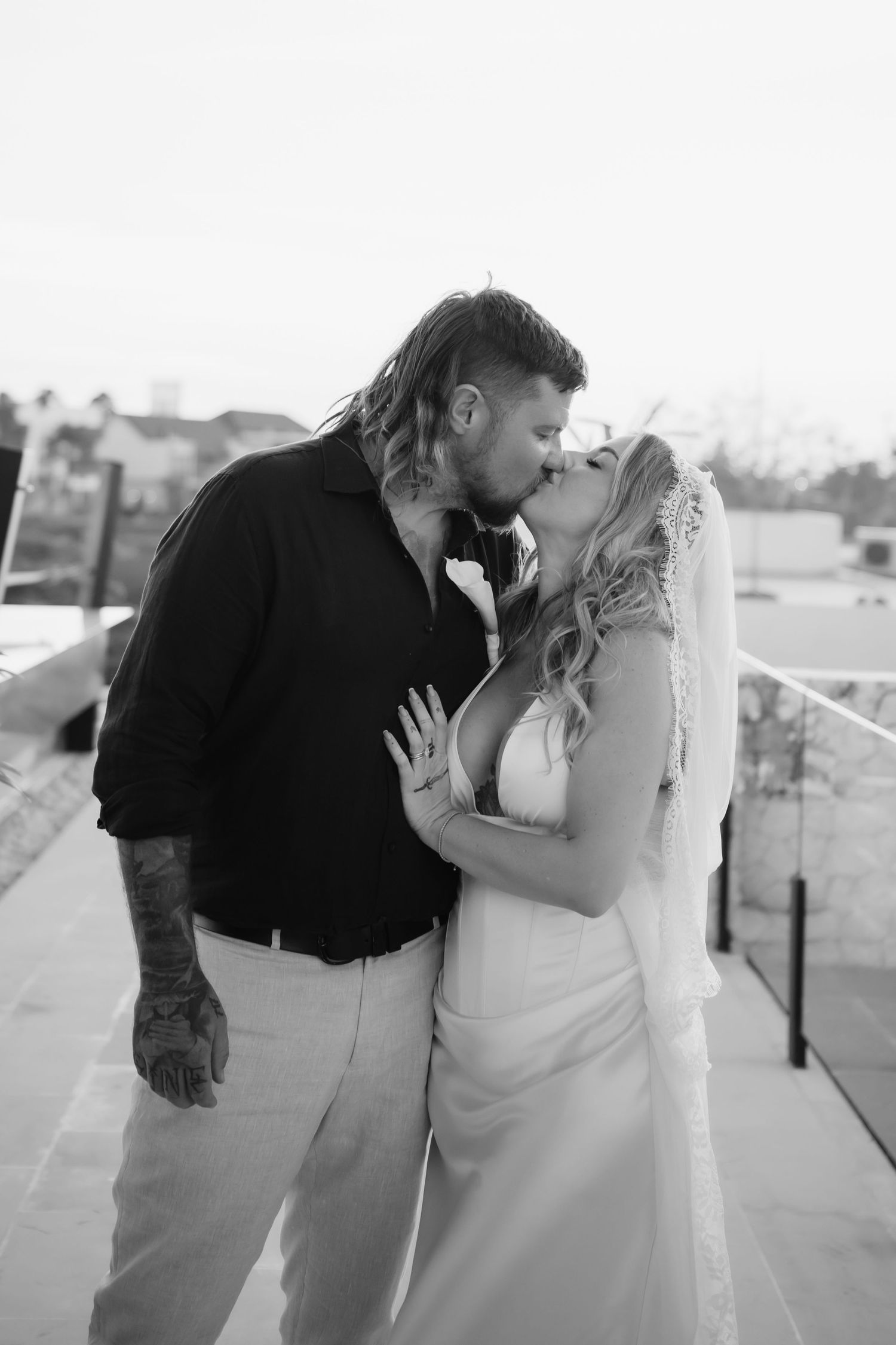 Black and white photo of tattooed groom kissing bride in white dress and veil on rooftop.