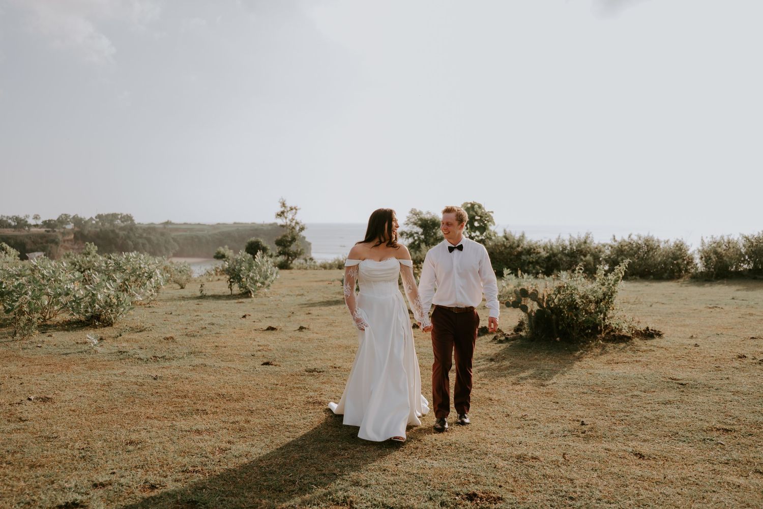 Bride and groom walk hand-in-hand through scenic coastal meadow on wedding day.