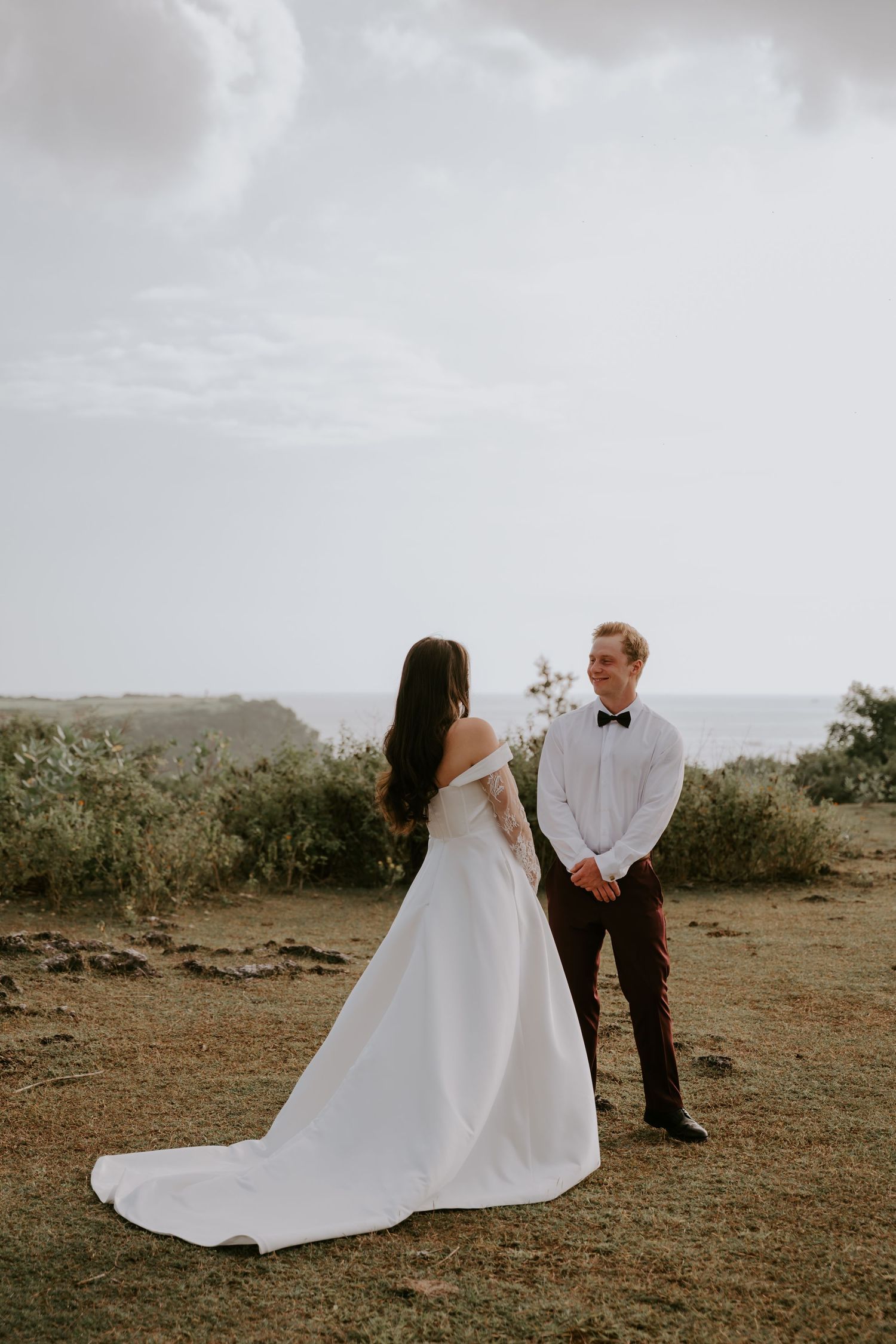 Bride in off-shoulder white gown with train faces groom in white shirt and bow tie at outdoor coastal wedding ceremony.