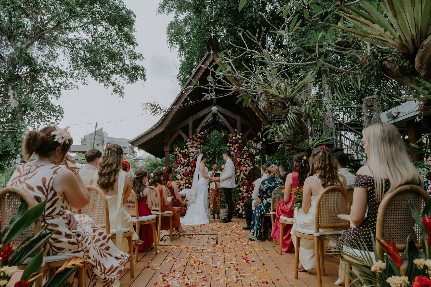 Tropical outdoor wedding ceremony with guests seated along a petal-lined aisle leading to a lush floral arch.