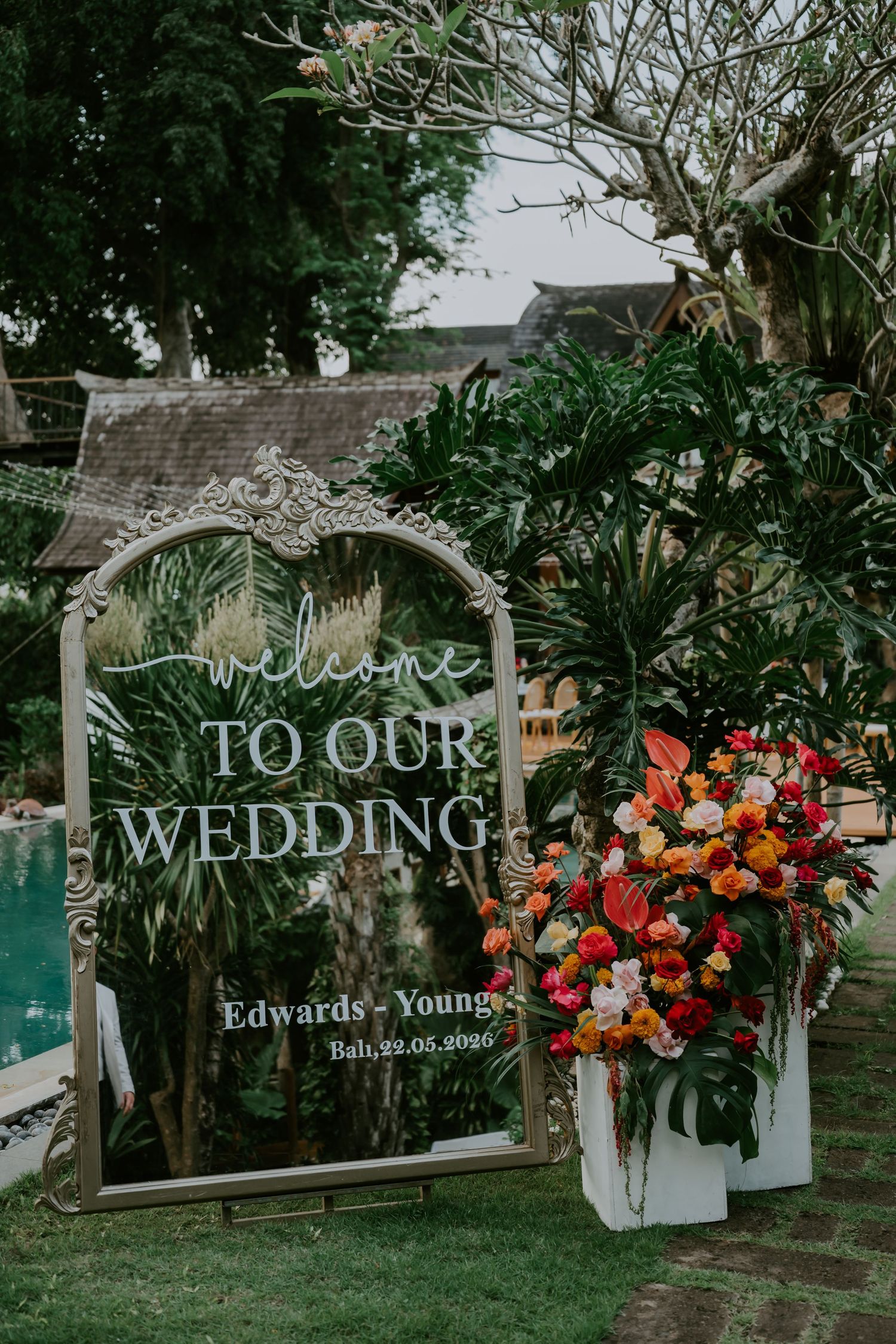 Ornate mirror welcome wedding sign for Edwards-Young Bali wedding surrounded by tropical floral arrangements.