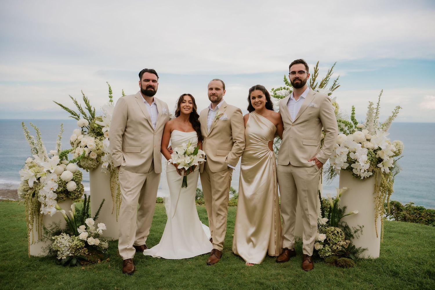 Wedding party of five posing in front of white floral arch with ocean backdrop on a grassy cliff.