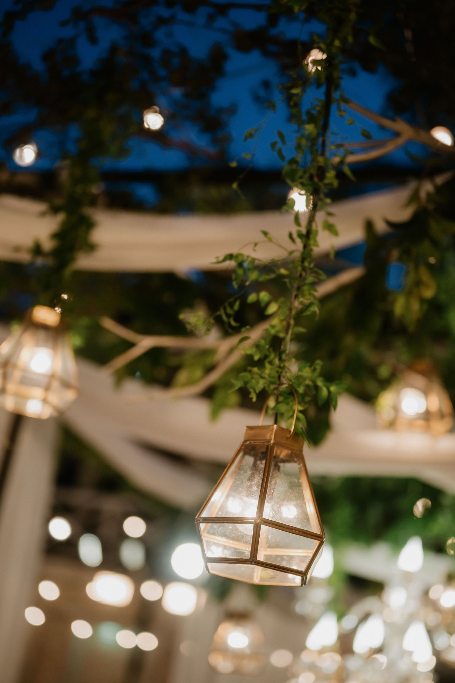 Geometric glass lantern hanging among greenery and string lights at outdoor evening wedding reception.