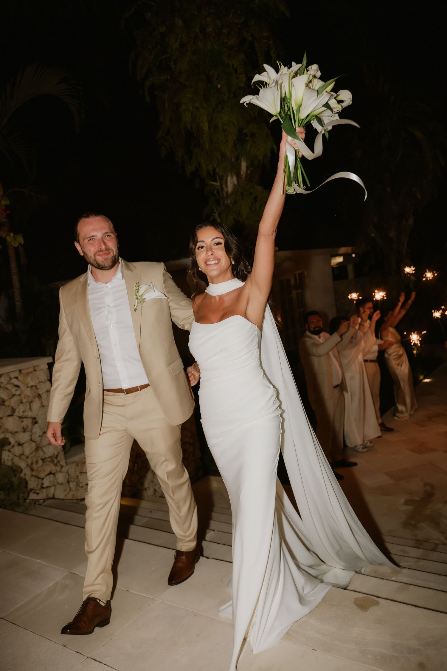 Bride raises calla lily bouquet triumphantly as couple exits wedding reception at night.