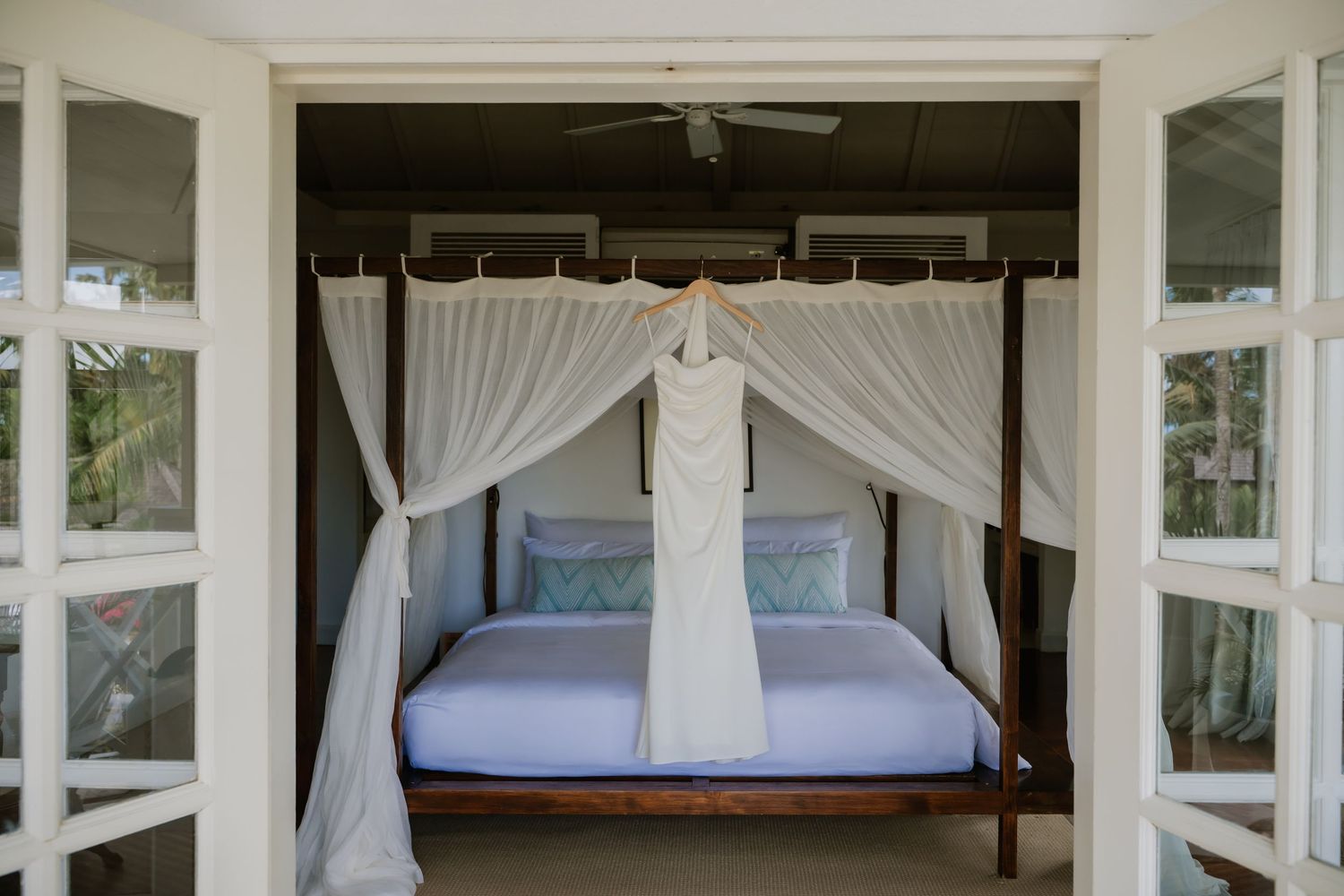 White wedding dress hanging on a canopy bed in an elegant tropical villa bedroom with white curtains.