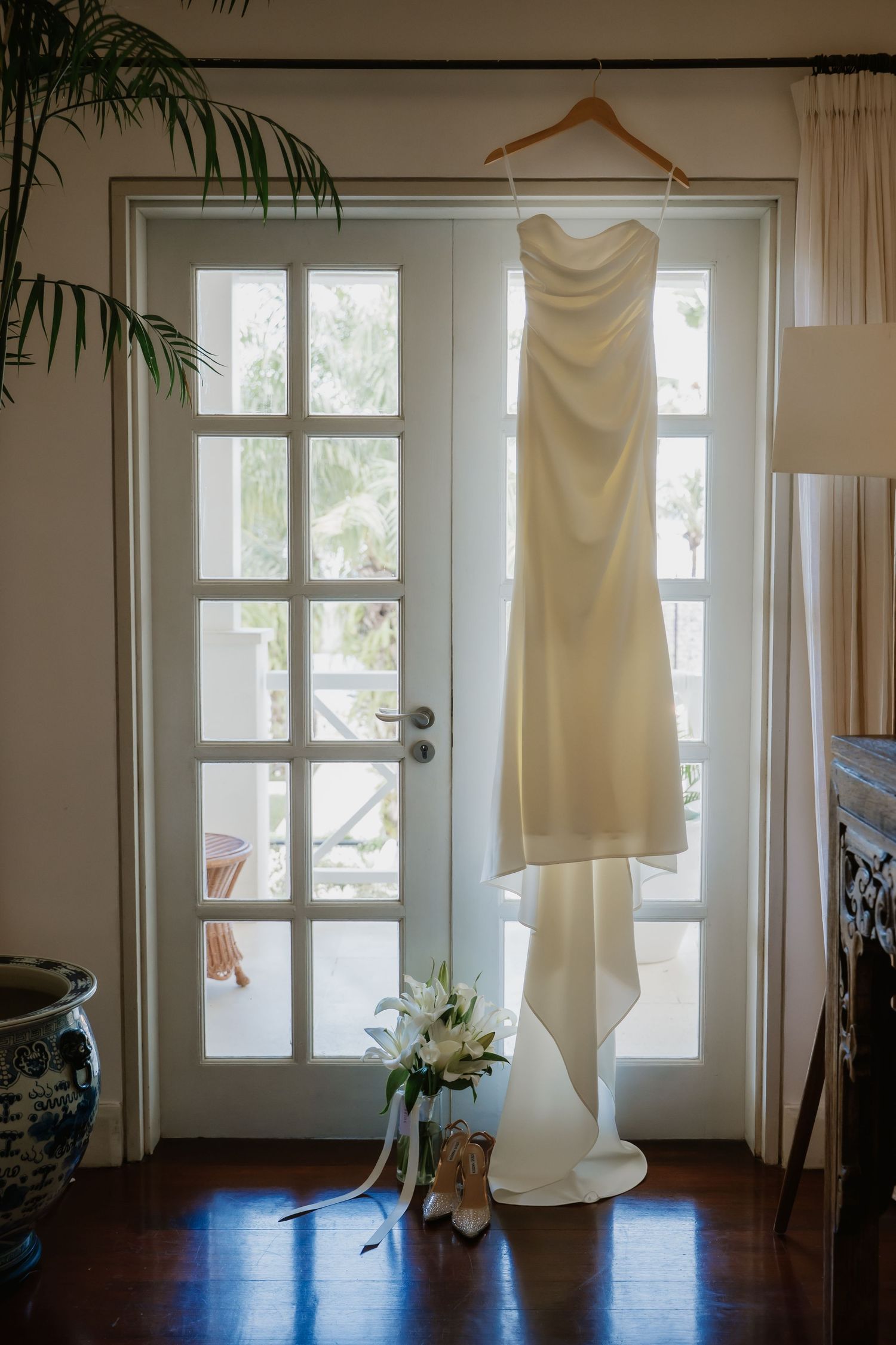 Elegant strapless white wedding gown hanging on French doors with bridal bouquet and heels.