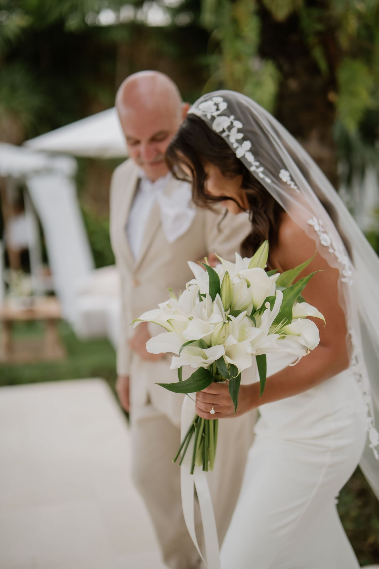 Bride in fitted white gown holding white lily bouquet walks with her father toward the outdoor ceremony.