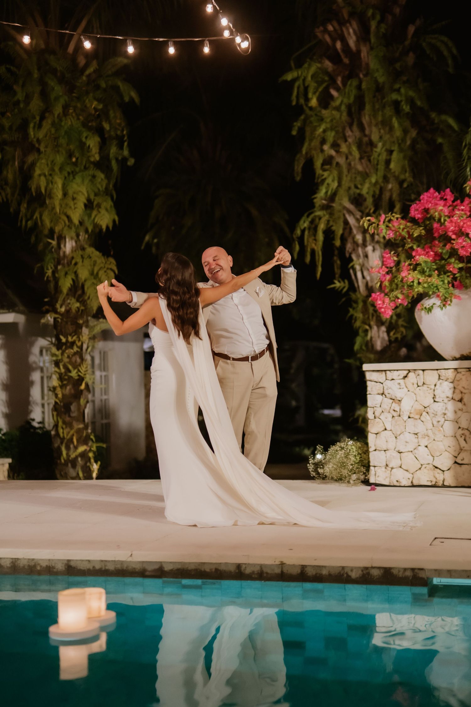 Bride and father share emotional dance beside illuminated pool surrounded by tropical night garden.
