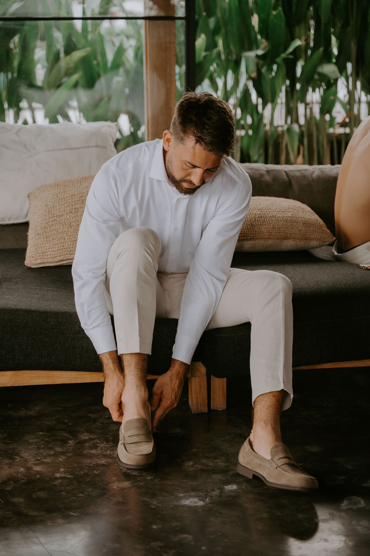 Groom in white linen suit putting on tan loafers while seated on couch with tropical backdrop.