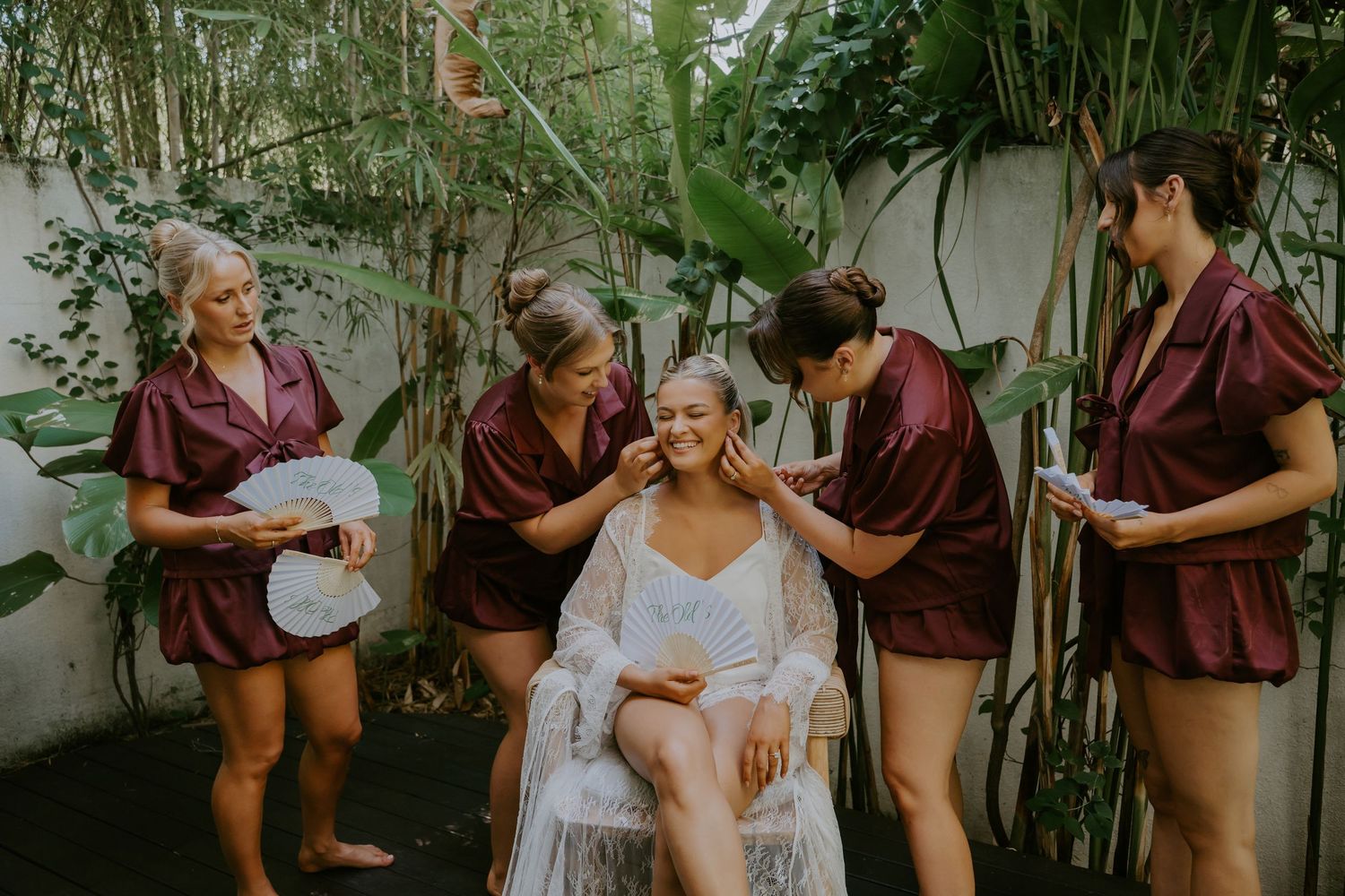 Bridesmaids in burgundy satin robes helping bride with makeup outdoors surrounded by tropical greenery.