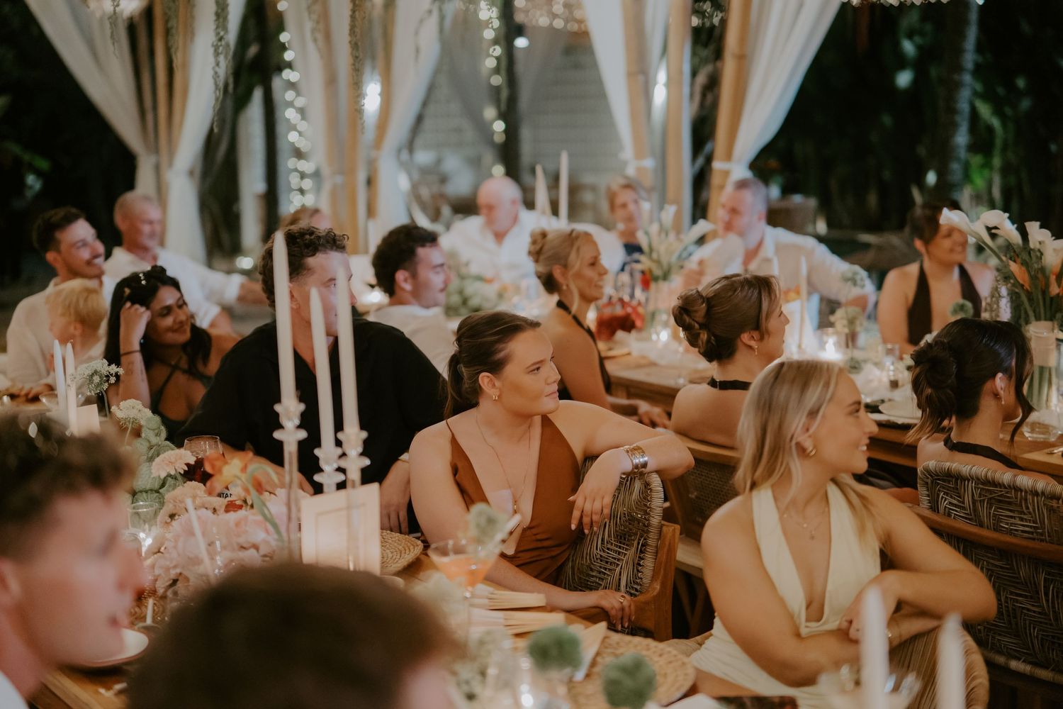 Wedding guests laugh and mingle at candlelit outdoor reception dinner under draped white curtains at night.