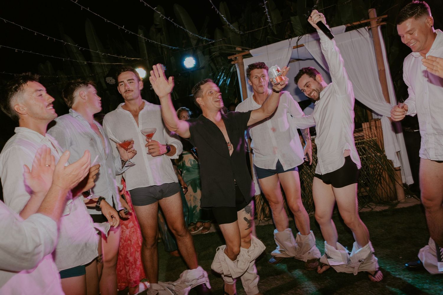 Groomsmen celebrate and dance energetically at lively tropical wedding reception party.