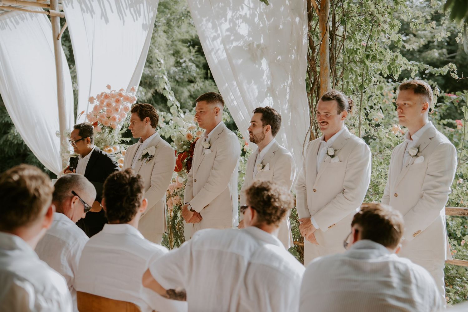 Groomsmen in white suits stand at outdoor wedding altar surrounded by lush greenery and floral decorations.