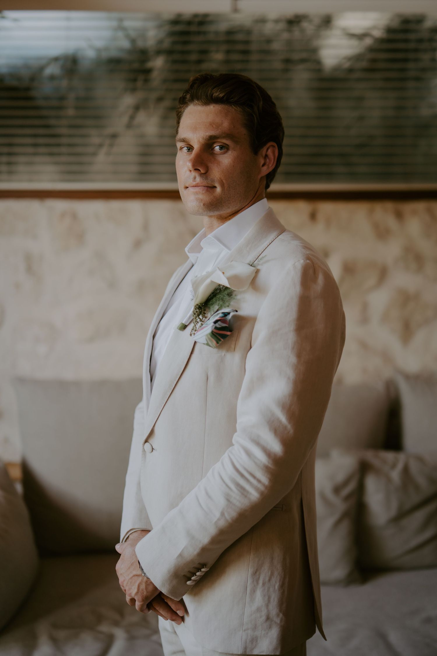Dapper groom in white linen suit with boutonniere poses confidently indoors before his wedding ceremony.