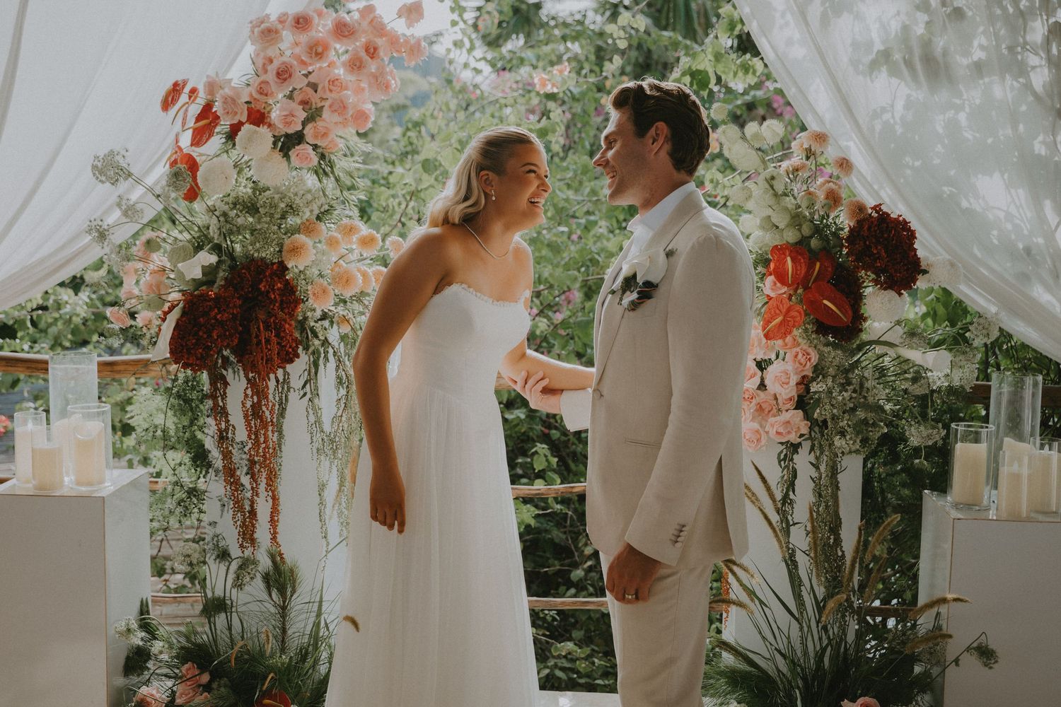 Bride and groom exchange vows at floral arch with pink and red blooms during outdoor wedding ceremony.