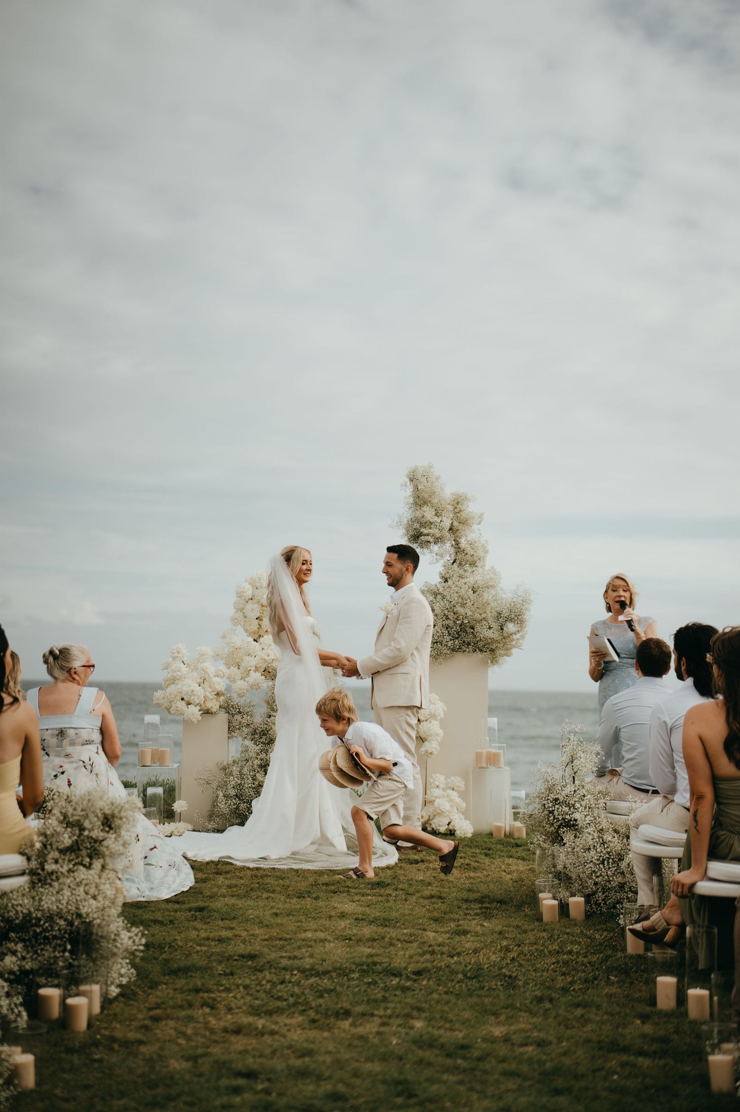Outdoor beachside wedding ceremony with bride and groom exchanging vows surrounded by white floral arrangements and guests.