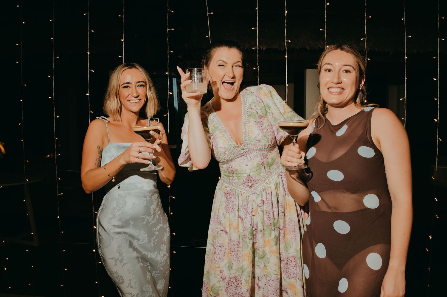 Three women in colorful dresses laugh and pose together at a wedding reception.