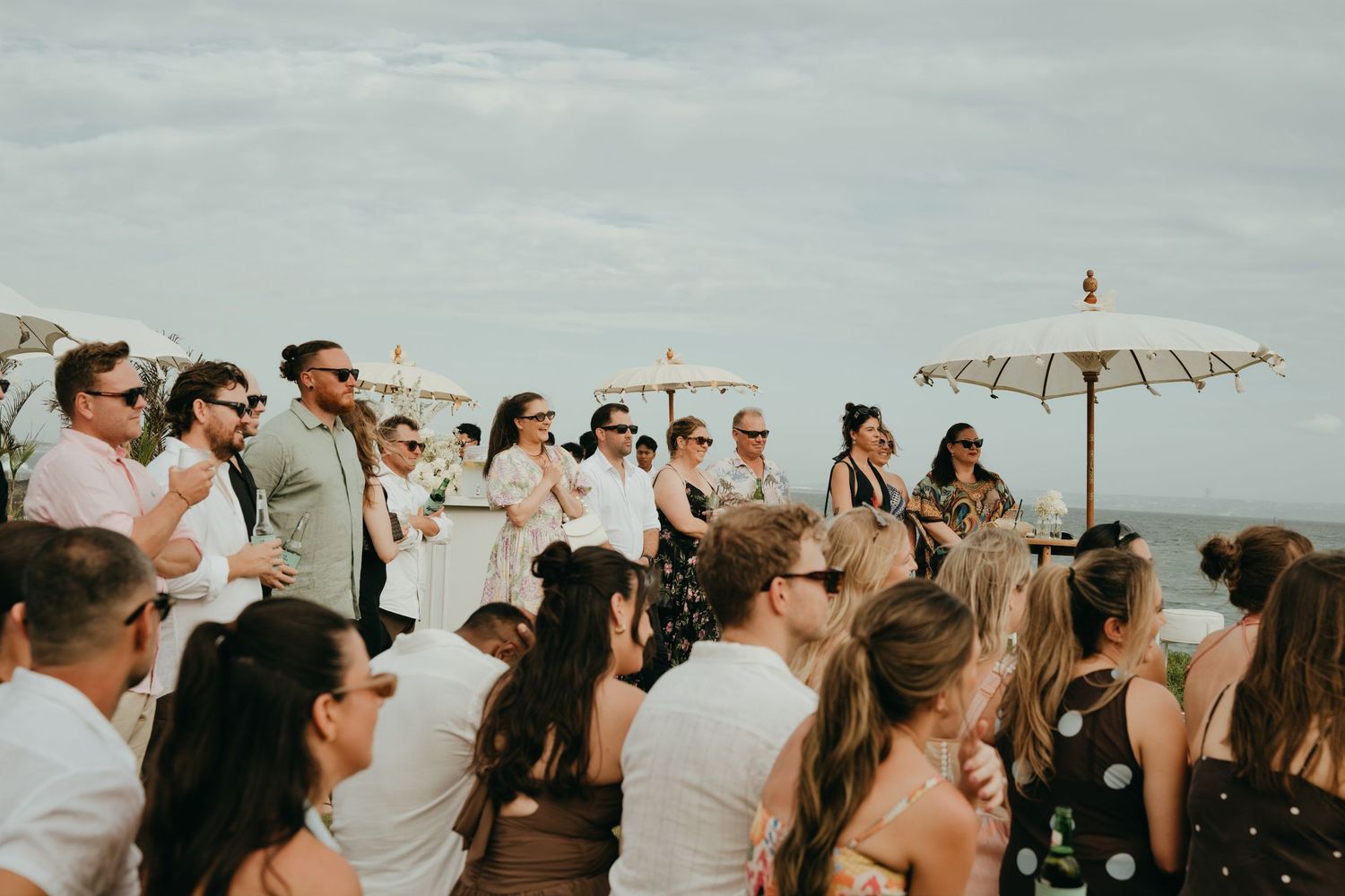Wedding guests seated outdoors under parasols with ocean view at tropical beach ceremony.