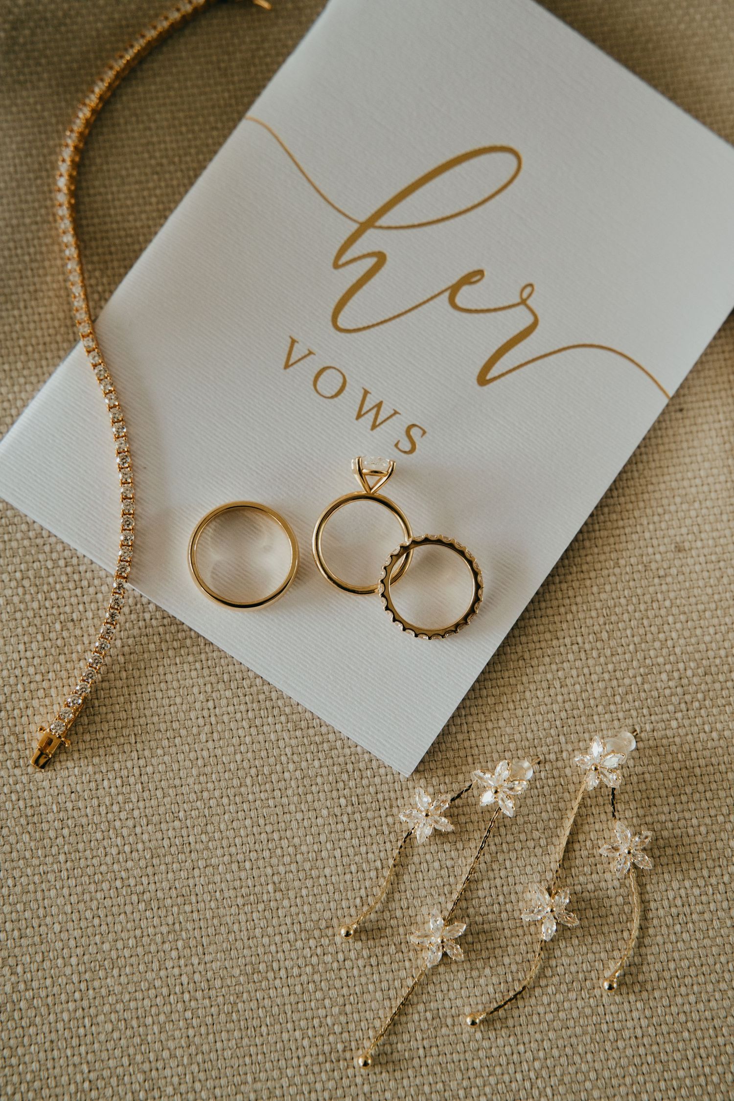 Gold wedding rings, floral earrings, and diamond necklace arranged on a white Her Vows card.