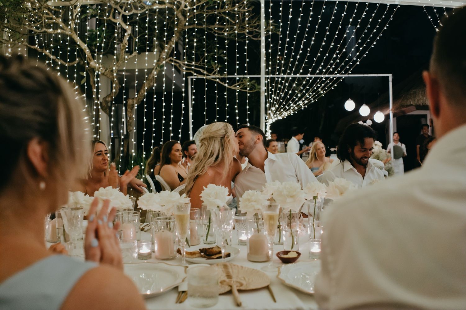 Wedding guests celebrate at a fairy light adorned reception dinner table at night.