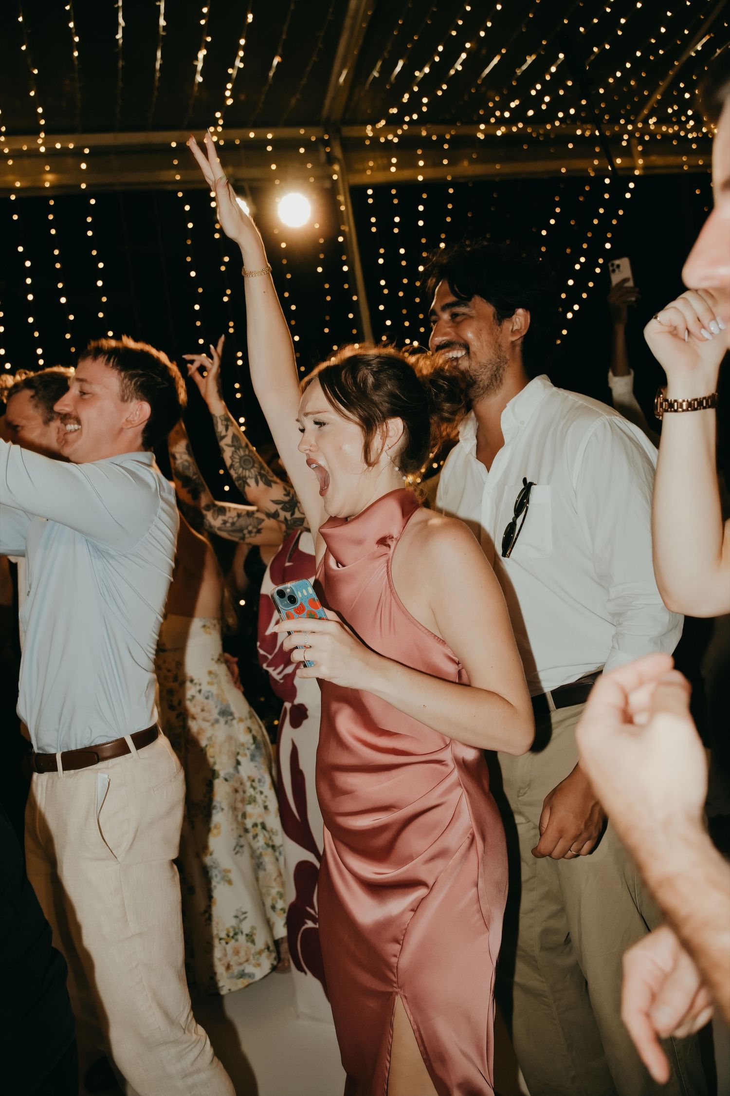 Wedding guest in a pink satin dress dances joyfully with arms raised under fairy lights at a reception.
