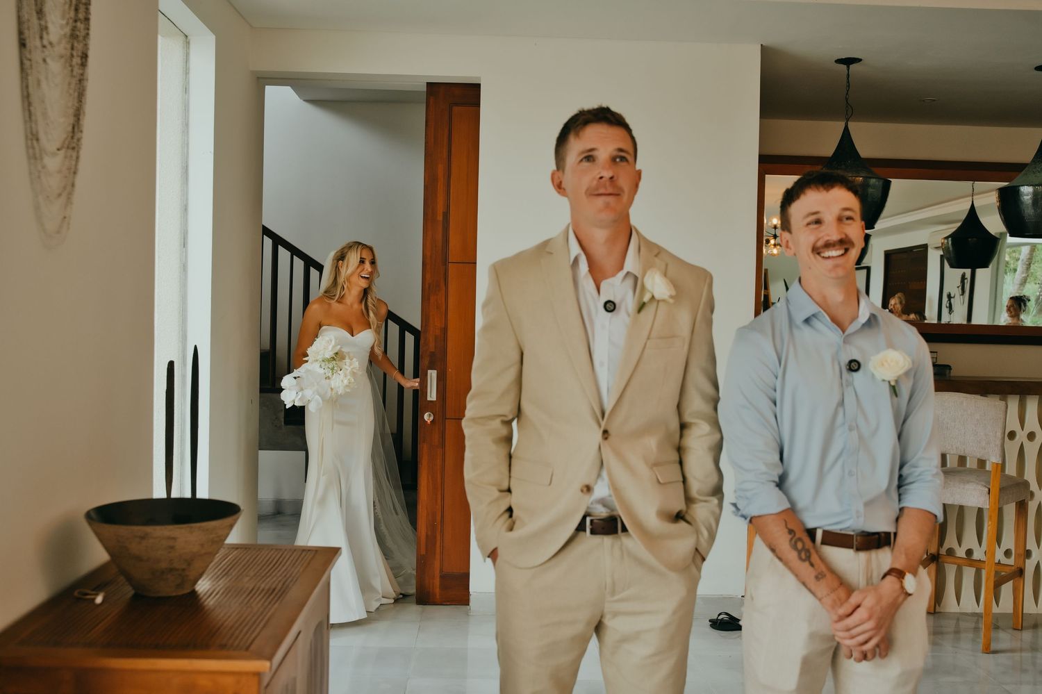 Groom in tan suit smiling as bride in white gown approaches during first look.