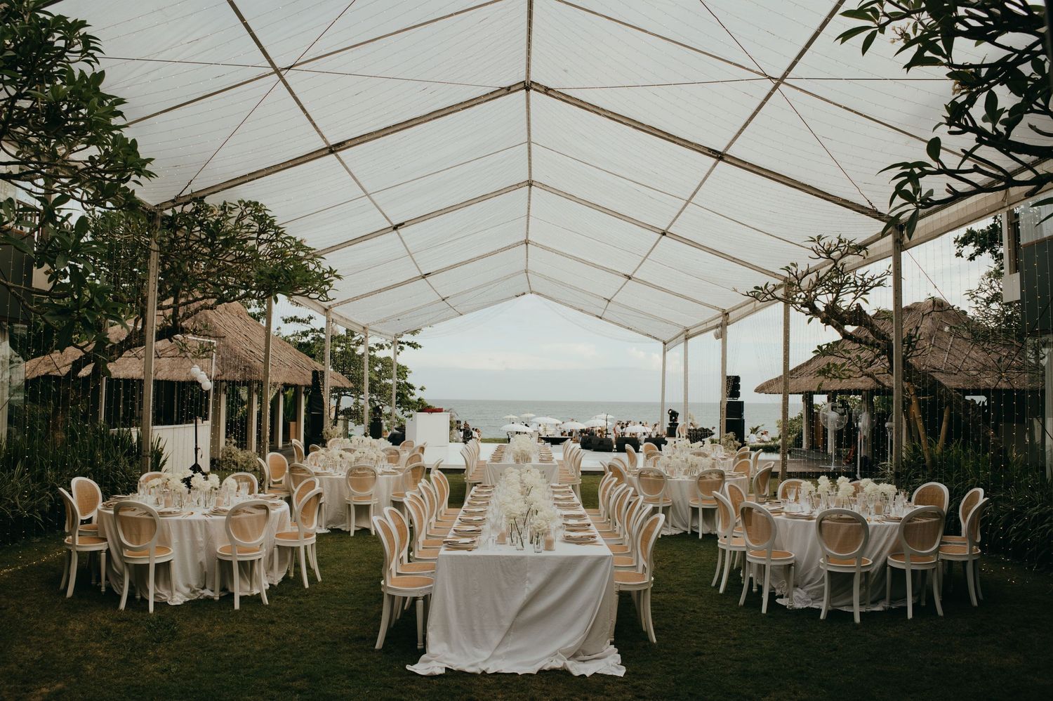 Elegant outdoor wedding reception under a white marquee tent with ocean views at dusk.